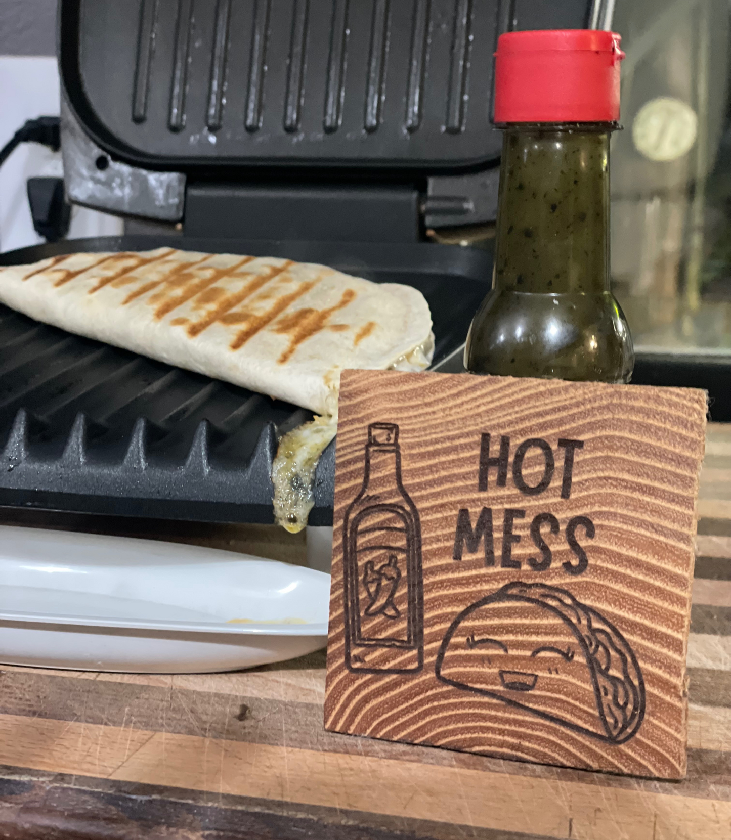 Features a wooden (oak) coaster with a laser-etched design showing a graphic of a smiling taco and a bottle  with two chili peppers on the label (depicting hot sauce), the coaster/tile also contains text reading "HOT MESS". The wooden tile is propped on a real bottle of hot sauce. The photo also shows a quesadilla on a grill in the background with cheese dripping down. 