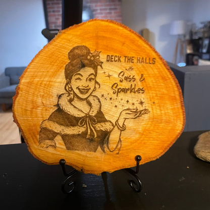 ~7 inch wooden plaque with engraved design of a girl with a mischievous grin holding her palm up towards levitating stars and text reading 'Deck the Halls with Sass & Sparkles', set on on a small metal easel stand. 