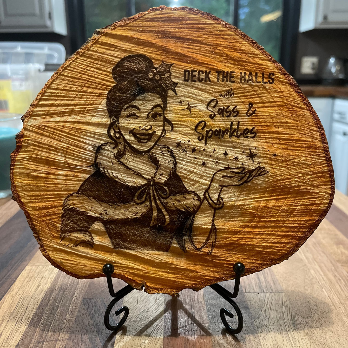~7 inch wooden plaque with engraved design of a girl with a mischievous grin holding her palm up towards levitating stars and text reading 'Deck the Halls with Sass & Sparkles', set on on a small metal easel stand.