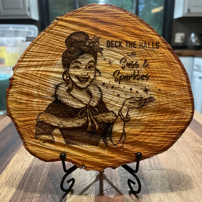 ~7 inch wooden plaque with engraved design of a girl with a mischievous grin holding her palm up towards levitating stars and text reading 'Deck the Halls with Sass & Sparkles', set on on a small metal easel stand.