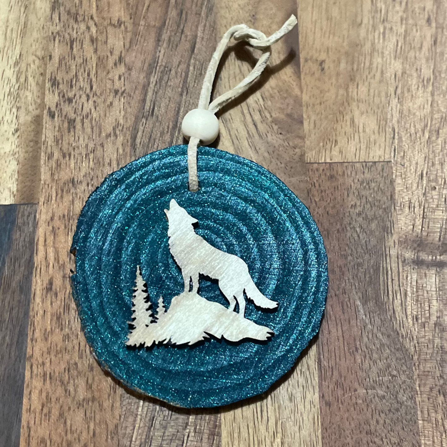 Wooden ornament with a wolf laser-cut design in a light stain on a wood round painted in dark metallic teal  Tied with a tan leather cord with a round wooden bead for embellishment.